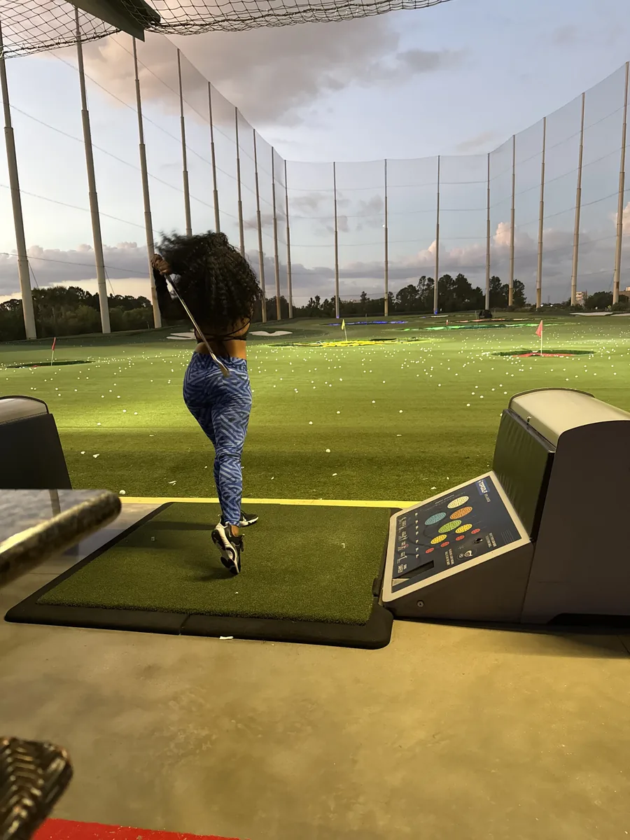 A golfer at the top of her follow-through in a Topgolf bay at dusk, the floodlit range and target greens stretching out beyond the netting