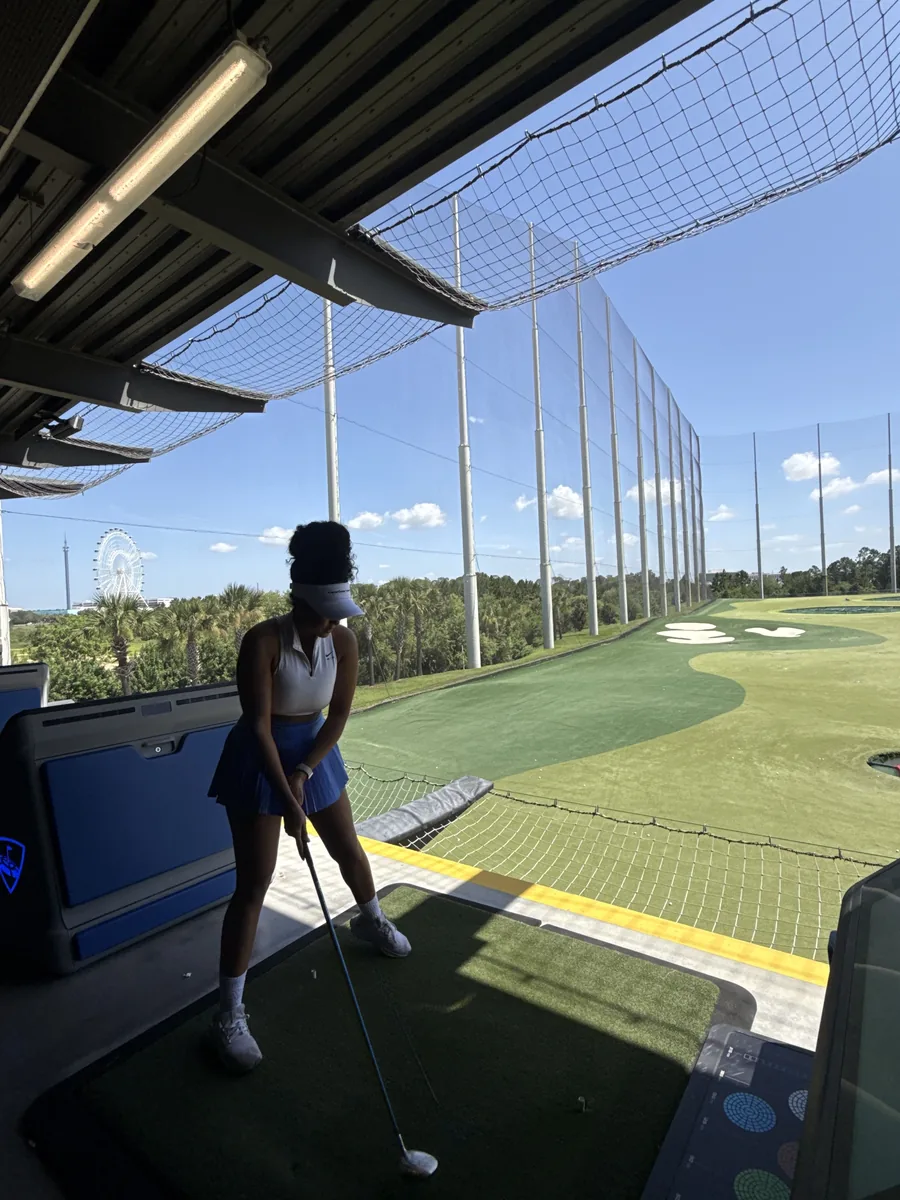 A golfer addressing the ball in a Topgolf bay on a bright afternoon, in a white polo, blue pleated skirt and visor, with the Orlando Eye on the skyline