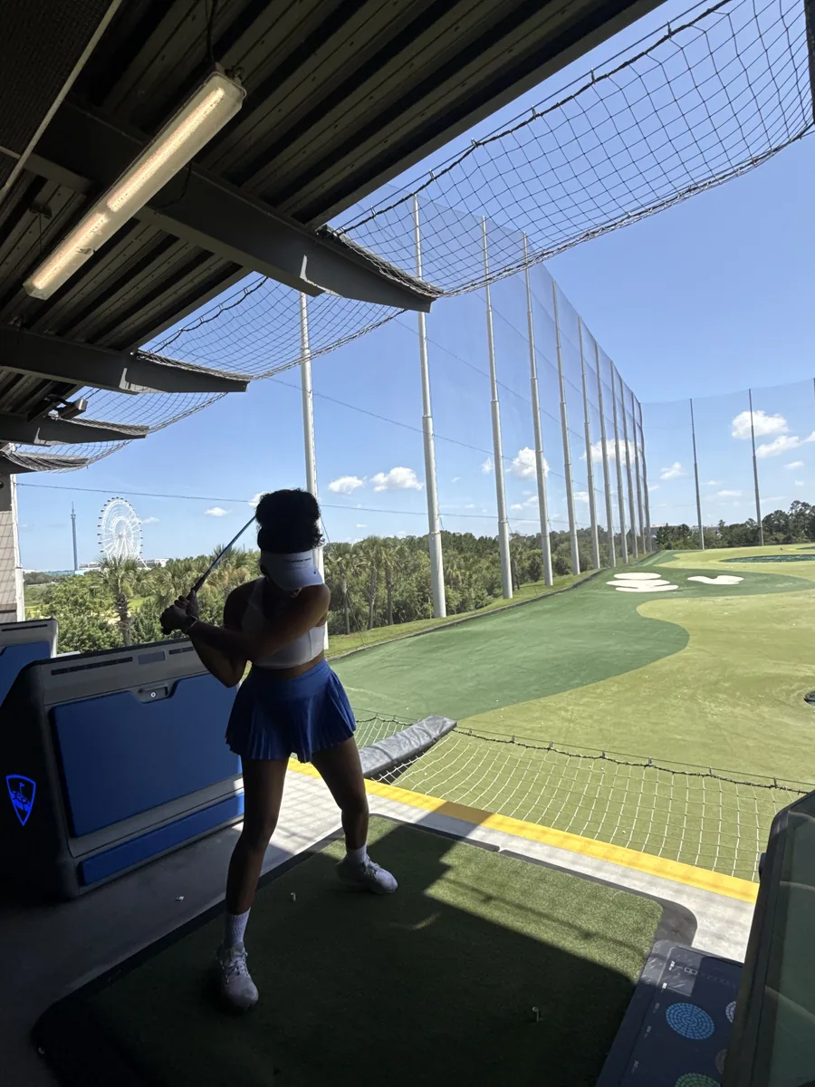 A golfer at the top of her backswing in a Topgolf bay, palm trees and the sweep of the range behind her