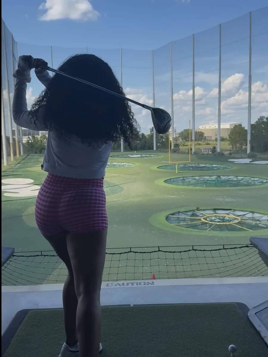 A golfer coiled at the top of her backswing, driver behind her shoulder, looking out over the target greens of the range