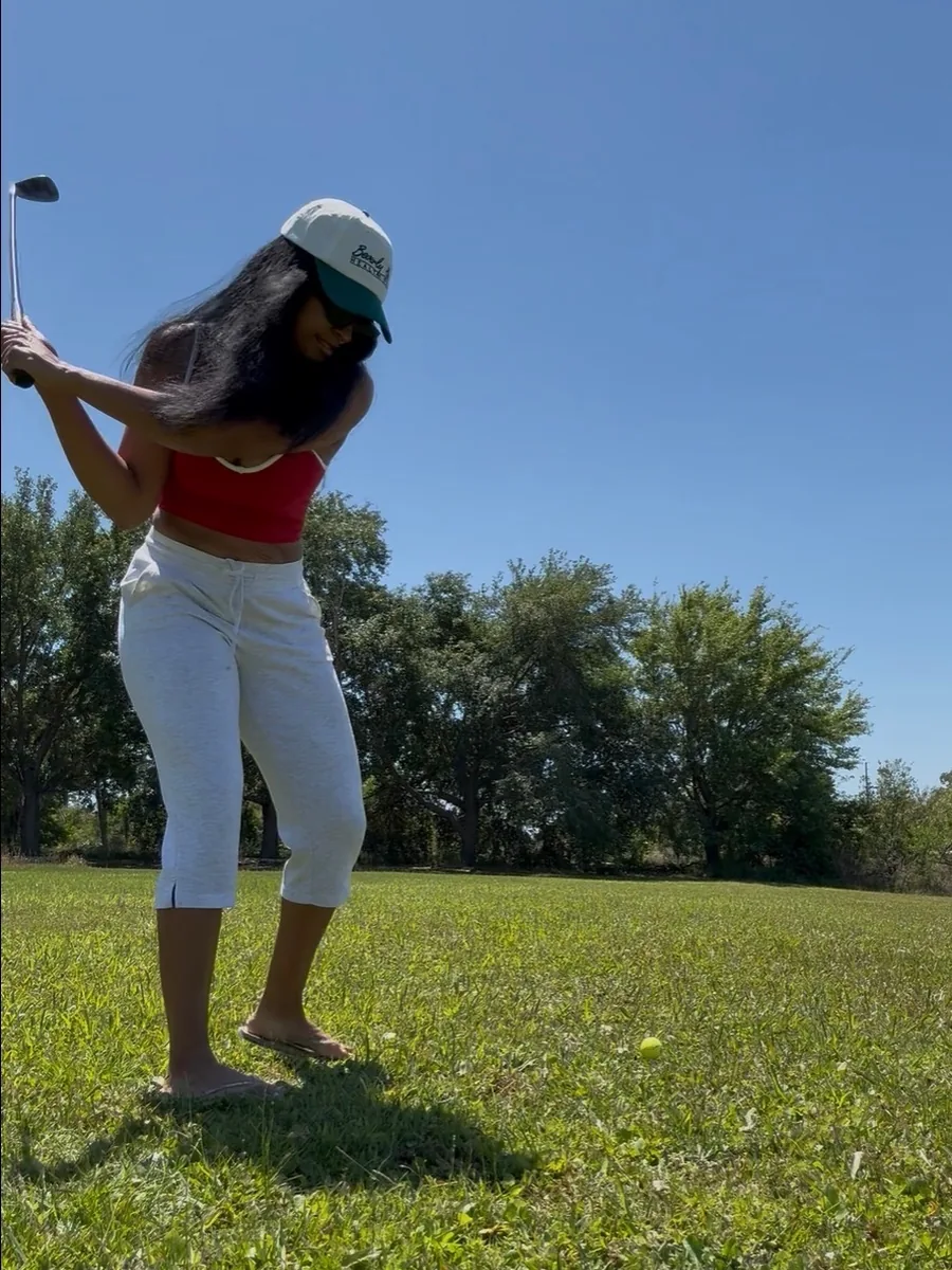 A golfer mid-swing on an open grass fairway under a clear blue sky, in a red top and white cap